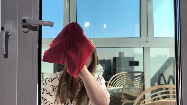 Young woman wipes glass door using cloth during sunny day. Female cleans window surface removing dust and marks manually. Spring cleaning routine highlights household care and hygiene..