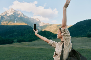 Naklejka premium Smiling woman in a beige jacket taking a selfie in a green meadow with mountains in the background, embodying outdoor lifestyle, travel and exploration adventure