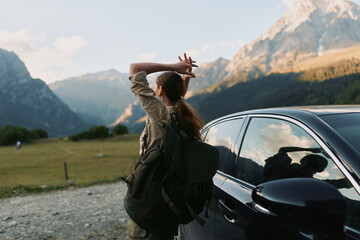 Fototapeta premium Young woman with a backpack stands beside a parked car in a valley, hands behind her head, gazing at distant mountains, enjoying a tranquil outdoor moment of freedom