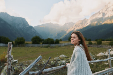 Naklejka premium Young woman in a cozy gray cardigan stands beside a weathered wooden fence in a mountain valley, smiling softly as distant peaks glow under a clear blue sky, conveying a peaceful outdoor lifestyle.