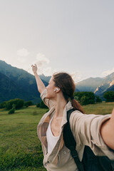 Naklejka premium Young woman with a backpack takes a selfie in a valley as mountains rise behind, her arm extended toward the camera with a joyful breeze.