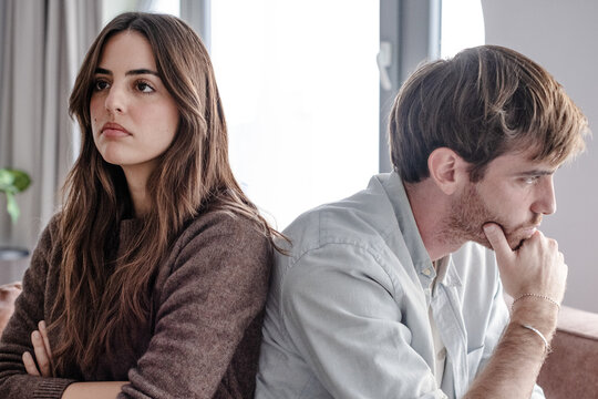 Young couple sitting back to back on couch having a dispute, expressing relationship conflict, unresolved problem or question, emotional distance, or communication problems in a modern living room.