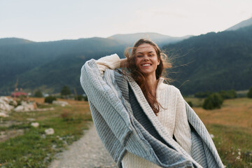 Fototapeta premium A happy woman in a white sweater and gray shawl smiles while standing on a rural path with mountains in the background. Lifestyle concept of freedom and nature.