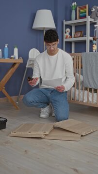 Man kneels holding drill and instruction sheet while assembling crib panels beside a baby crib in building; new parent frustration.