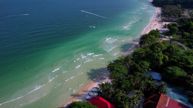 Aerial video of the amazing emerald water white sand Sao Beach with lively water sports, tourists, cabanas and sun umbrellas, beach bar and parasailing. Hot sunny day with long waves on Phu Quoc Islan