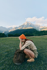 Naklejka premium Young woman crouches in a sunlit meadow with a backpack, shielding her eyes as rugged mountains rise in the background.
