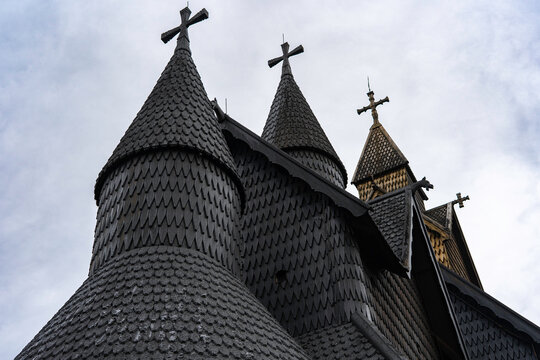 Traditional Norwegian stave church surrounded by spring countryside and forested hills under dramatic cloudy sky in Telemark, Norway. Dark wooden medieval-style architecture. 