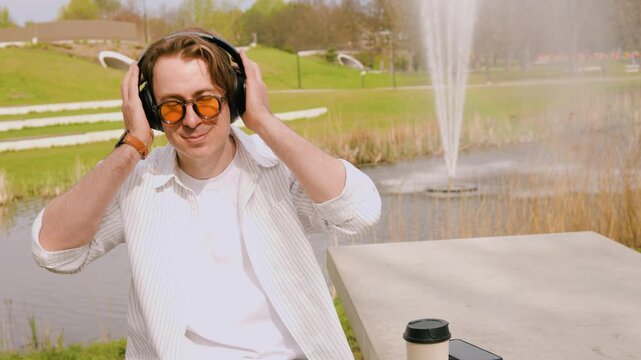 A young man in orange sunglasses enjoys music through headphones in a city park. 4K video featuring a sunny day, park fountain, and outdoor relaxation. High quality footage