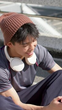 Vertical video: Starting clip, Asian man checking phone on steps, listening with beanie headphones