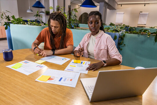 Diverse coworkers sitting at communal wooden table in coworking space reviewing charts and laptop