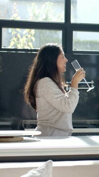 Vertical video: Holding flute woman in sweatshirt raising glass, miming and sipping at kitchen