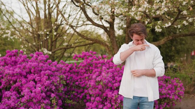 A man suffers from allergy season and hay fever in a park with spring blooms. 4K video showing sneezing and itchy eyes caused by high pollen levels from flowers. High quality footage