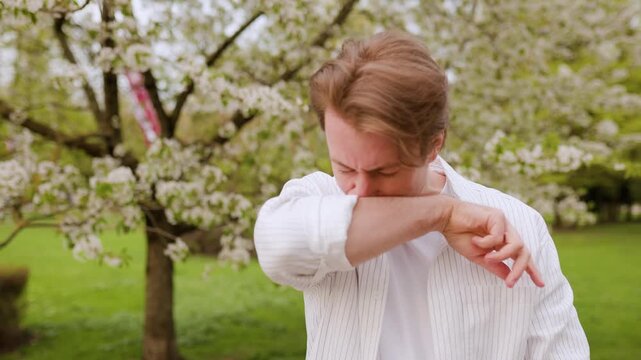 Man struggling with allergy season and hay fever amid spring blooms. 4K footage of sneezing and itchy eyes caused by pollen in a scenic park. High quality