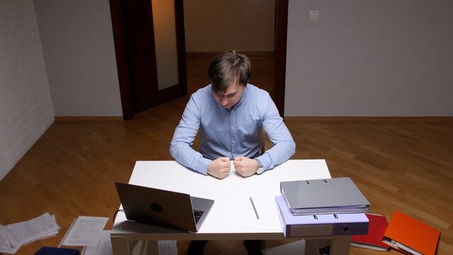 Man with clenched fists on a desk showing his work frustration and stress, experiencing a professional failure in an office setting with documents and a laptop