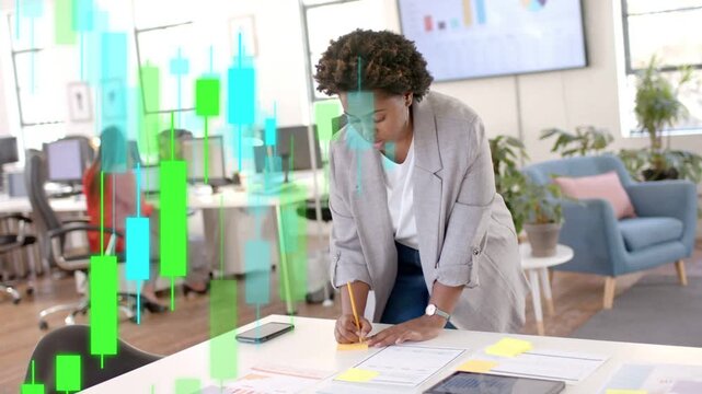 Woman leaning over writing notes for finance planning, charts rising from left moving across desk