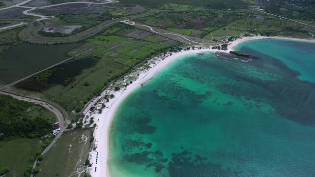 Aerial View Emerald Lagoon With Curved White Beach, Adjacent Farmland And Green Hills, Clear Shallow Patches And Reef Patterns, Calm Atmosphere Suited For Nature And Travel Visuals,Lombok