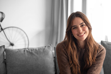 Naklejka premium Smiling young woman sitting on a sofa in a cozy living room, wearing a casual sweater and looking relaxed and approachable; natural light and warm tones create a comfortable, friendly atmosphere.