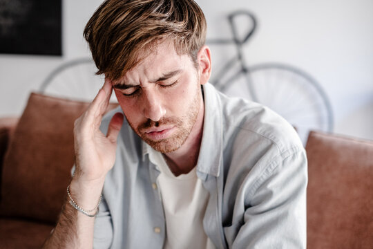 A young man sits on a couch at home holding his temple and wincing from a headache. The image conveys stress, fatigue and discomfort in a quiet domestic living room setting.