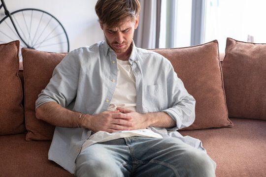 A young man sits on a living room couch clutching his lower abdomen and wincing in visible discomfort, suggesting stomach ache, indigestion or stress-related abdominal pain while at home.