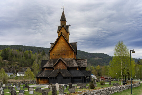 Traditional Norwegian stave church surrounded by spring countryside and forested hills under dramatic cloudy sky in Telemark, Norway. Dark wooden medieval-style architecture.