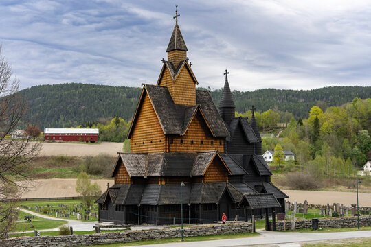 Traditional Norwegian stave church surrounded by spring countryside and forested hills under dramatic cloudy sky in Telemark, Norway. Dark wooden medieval-style architecture.