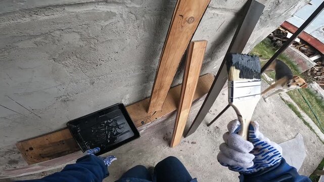 The process of painting wood. A builder paints a wooden beam black. A worker in a blue uniform and protective gloves applies paint with a brush. First-person view (POV). Redesign, handmade