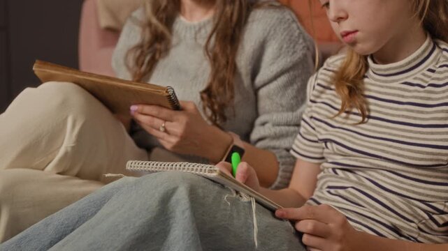 Cropped shot of preteen girl drawing in sketchbook with felt tip pen sitting next to mother while spending quality time with parent during weekend at home