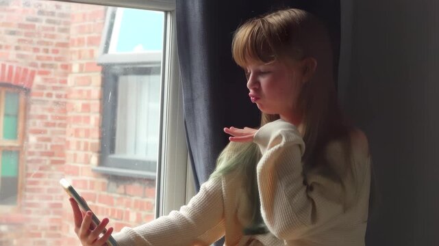 A girl sitting with a smartphone by a window. Making funny faces and fooling around while talking to friends.