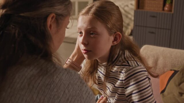 Over shoulder shot of adult mother tucking daughters hair behind ears then kissing preteen girl on forehead while comforting upset child in living room