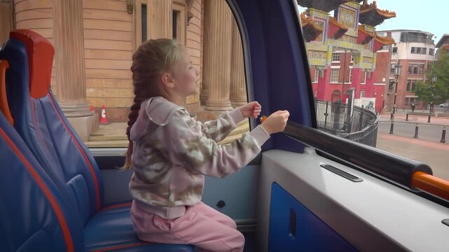 A girl sits on the upper deck of a city bus near the front windshield. The bus stops at a traffic light. She plays pretending her hands hold a steering wheel. Liverpool suburbs view
