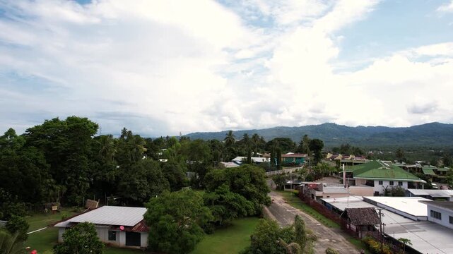 Aerial view of lush tropical vegetation, residential houses with white roofs, and distant mountains under a cloudy sky in Wewak, East Sepik Province, Papua New Guinea.