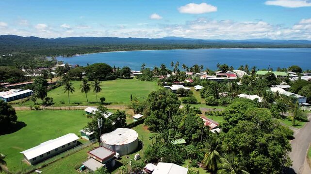 Aerial view of the coastal town featuring lush tropical vegetation, residential houses, and a calm blue bay under a bright sky in Wewak, East Sepik Province, Papua New Guinea.
