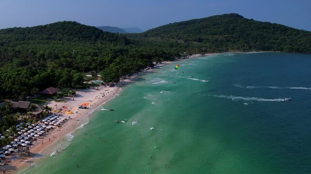 Aerial video of the amazing emerald water white sand Sao Beach with lively water sports, tourists, cabanas and sun umbrellas, beach bar and parasailing. Hot sunny day with long waves on Phu Quoc Islan