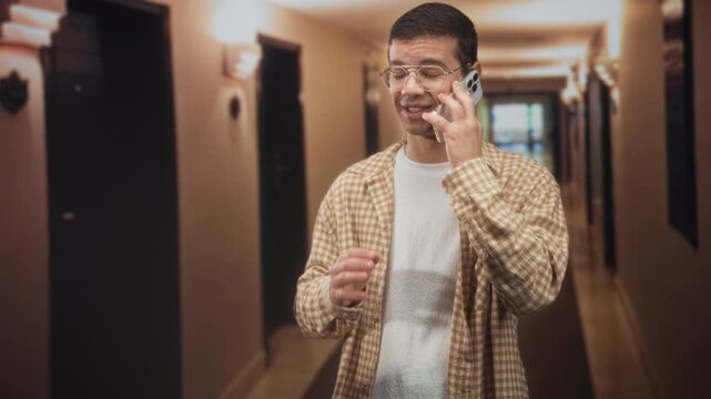 Young man in brown plaid shirt holds smartphone to ear and gestures with his hand while standing in a hotel corridor building; private call concern.