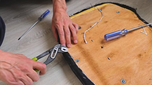 Hands of a person repairing an old office chair by removing staples from the fabric and securing the upholstery with a screwdriver on a wooden seat base