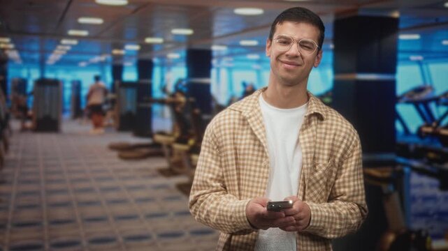 Man holding smartphone, tapping the screen while smiling and looking down in a building gym with treadmills and weight machines; quiet contentment.