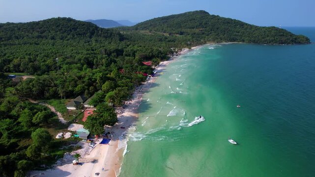 Aerial video of the amazing emerald water white sand Sao Beach with lively water sports, tourists, cabanas and sun umbrellas, beach bar and parasailing. Hot sunny day with long waves on Phu Quoc Islan