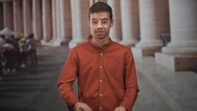 Man with glasses rubbing his eyes in a stone colonnade building, wearing rust shirt and clear plastic frames; fatigue introspection.