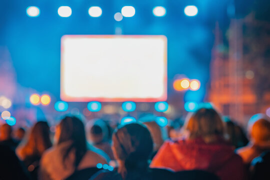 Rear view of blurred crowd watches large screen at night outdoor event with lights, bokeh, people silhouettes. Concept of screen advertising, cinema, festival, presentation, entertainment, copy space