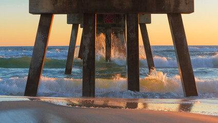 Pier structure stands tall over ocean waves at sunset, creating a serene atmosphere with reflections on wet sand and vibrant colors in the sky © JuliaDorian