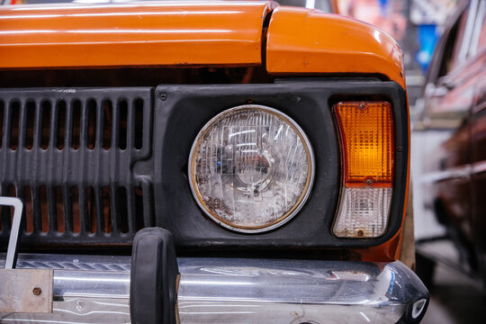 Detailed close-up captures front right corner of vintage orange car. Focus on round headlight, ribbed grille, and amber turn signal against vibrant paintwork, evoking nostalgia.