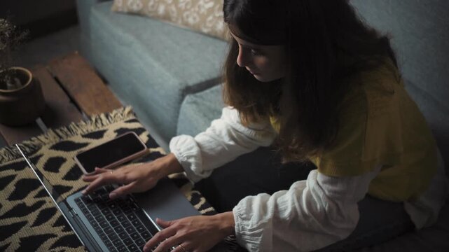 Young woman typing on her laptop while sitting on the sofa at home. Remote work and digital lifestyle concept, video clip.