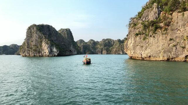 Small traditional wooden boat sailing through limestone karsts in Halong Bay Vietnam during golden hour sunlight