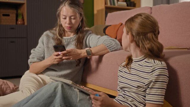 Rack focus of preteen girl drawing in sketchbook and talking to mom while woman texting on smartphone distracted by social media during shared leisure time in living room, disengaged parenting moment