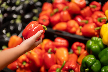 Woman hand choosing fresh red bell pepper in supermarket produce section, colorful vegetables...