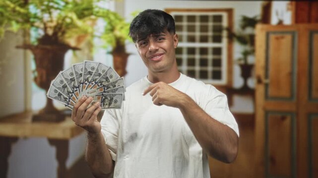 Young hispanic man holding a fan of hundred dollar bills and rubbing his eye while posing inside a building with plants nearby; playful bravado.