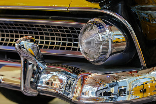Detailed close-up captures front end of vintage car, showcasing polished chrome bumper, checkered grille, and glass headlight with chrome accents. Reflective surfaces mirror classic automobiles and