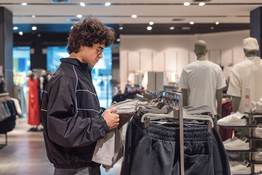 Young teenage boy with curly hair browsing shorts selection at modern menswear clothing store. Gen Z shopping behavior and consumer independence, teen fashion choices and personal style development