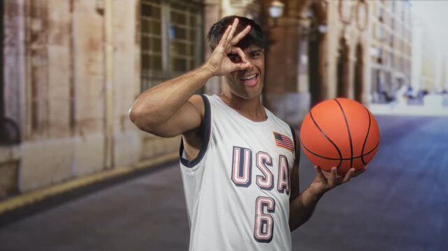 Man in usa basketball uniform holds an orange basketball and makes ok sign with his hand on a street in front of a building; playful confidence.