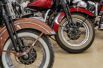 Obraz premium Detailed view captures front wheels and fenders of two vintage motorcycles parked indoors. Foreground motorcycle features a copper fender and whitewall tire, while background motorcycle showcases a
